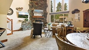 A cozy living room with a stone fireplace, a large window with a view, and a dining area with a round table. - Luna Ridge Chalet + Hot Tub + Mins to Bear Mtn (Big Bear Lake)