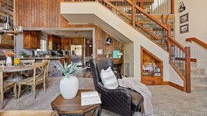 A spacious living area with a large wooden staircase, a dining table with chairs, and a cozy seating area with a chair and a side table. - Luna Ridge Chalet + Hot Tub + Mins to Bear Mtn (Big Bear Lake)