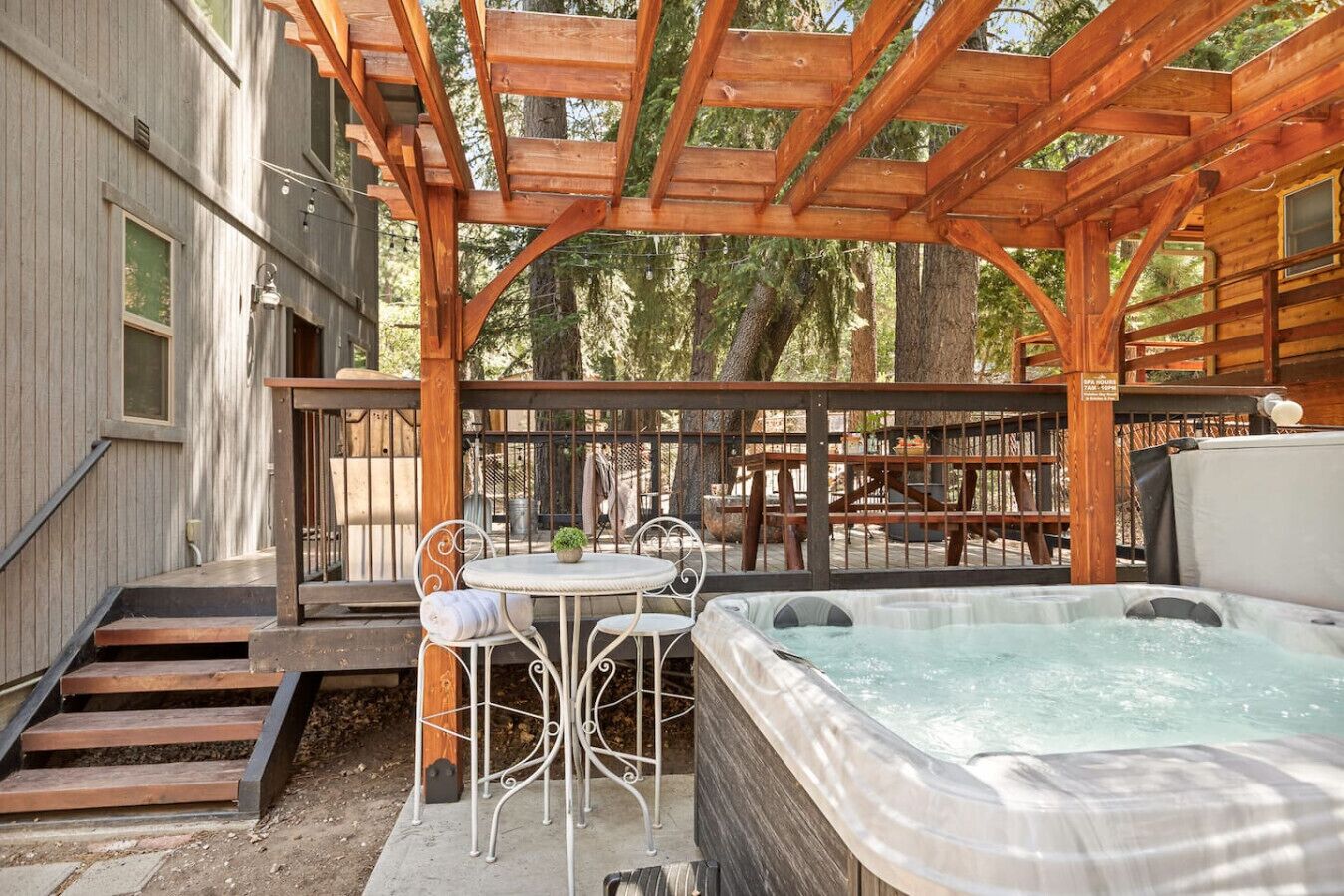 A wooden deck with a hot tub, a table with chairs, and a staircase leading to a building.