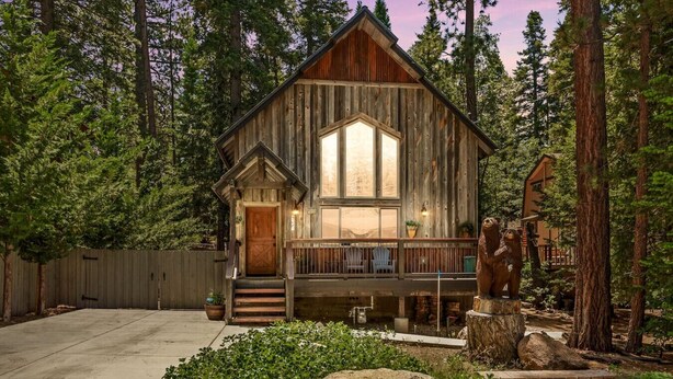 A rustic cabin with a wooden exterior, large windows, a porch with chairs, and a bear statue in the yard. - Luna Ridge Chalet + Hot Tub + Mins to Bear Mtn (Big Bear Lake)
