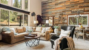 A cozy living room with a large window, a wooden wall, a 'welcome to our home' sign, and a dining area with a table and chairs. - Luna Ridge Chalet + Hot Tub + Mins to Bear Mtn (Big Bear Lake)