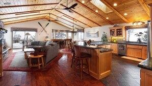 A spacious living area with a fireplace, a dining area, and a kitchen with wooden cabinets and a bar. - Treetop Oasis| Hot Tub, Lake Views, AC & Game Room (Big Bear Lake)