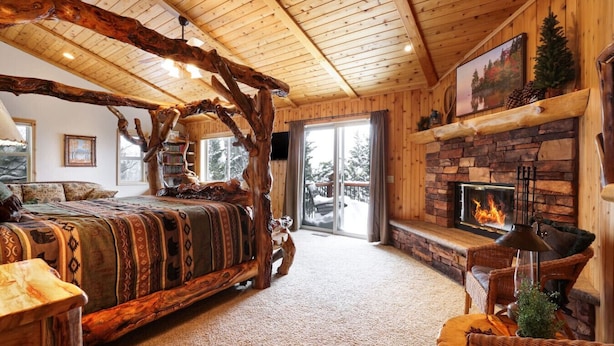 A log cabin interior with a rustic bed, a stone fireplace, and a balcony with a view of trees. - Treetop Oasis| Hot Tub, Lake Views, AC & Game Room (Big Bear Lake)