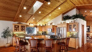 A spacious kitchen with wooden cabinets, a large island with bar stools, and a view of the outdoors. - Treetop Oasis| Hot Tub, Lake Views, AC & Game Room (Big Bear Lake)