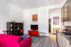 Ein gemütlicher Wohnraum mit rotem Sofa, Bücherregal, Fernseher und einer Küchenzeile.