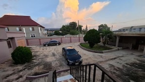 A residential area with a paved driveway, two parked cars, a staircase leading to a gated entrance, and a large building with a red roof in the background. - Rankbel Holidays Apartment (Kumasi)