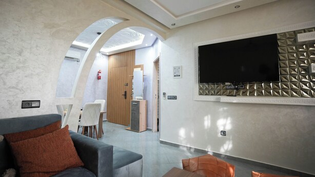 Ein modernes Wohnzimmer mit grauem Sofa, orangefarbenen Kissen, einem flachbildfernseher an einer strukturierten Wand und einem Essbereich mit Holztür und weißen Stühlen.