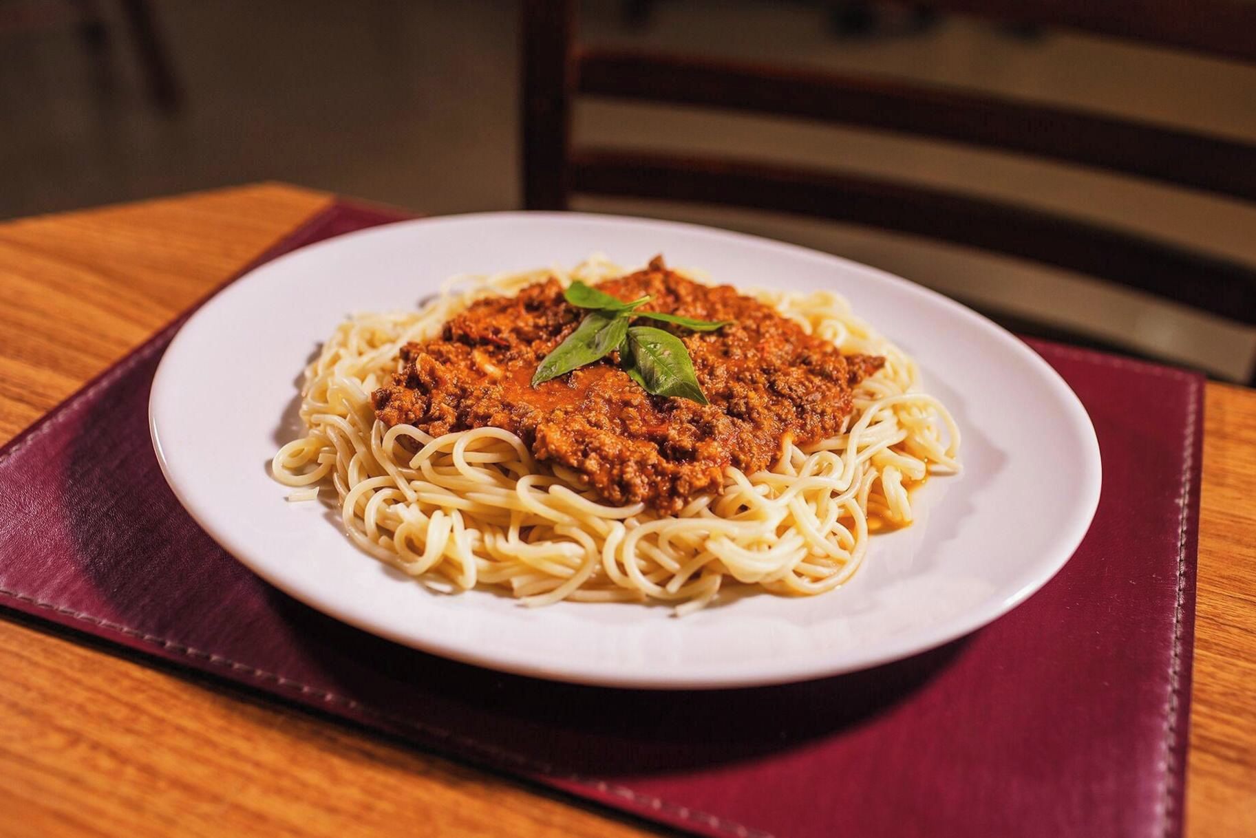 Um prato de espaguete com molho de carne e manjericão, sobre um prato branco, apoiado em uma mesa de madeira com um guardanapo vermelho por baixo.