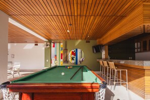 Una mesa de billar con superficie de fieltro verde, rodeada de sillas de mimbre, en una habitación con vigas de madera en el techo y una pared de acento verde.