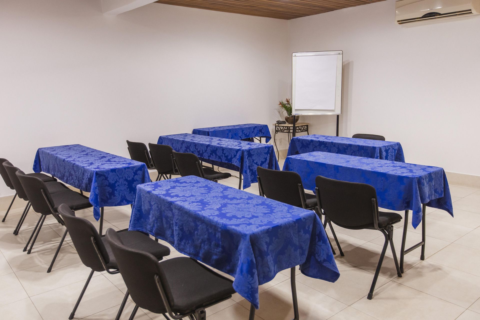 Uma sala de conferências com várias mesas cobertas por toalhas azuis, cadeiras pretas e um quadro branco na parede.