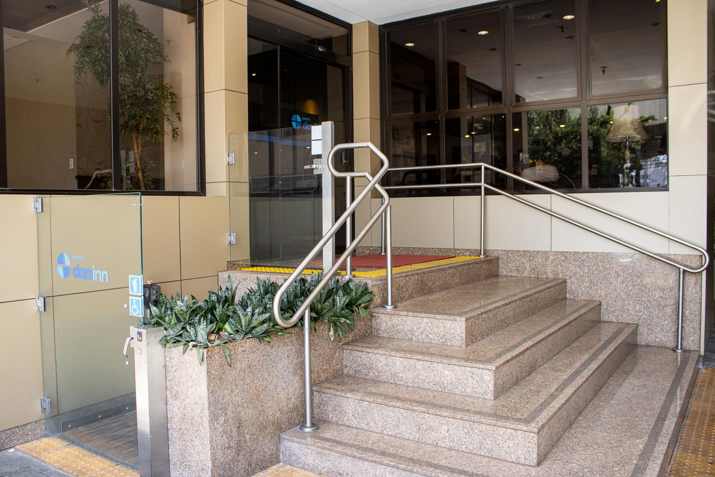 Entrada de hotel com porta de vidro, corrimão de metal e rampa de acesso.