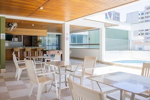 A modern indoor space with a dining area featuring white chairs and tables, a kitchen area with wooden cabinets, and large windows providing views of the outside.