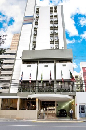 Un edificio moderno con varias banderas, un acceso cubierto y un coche estacionado en la entrada.