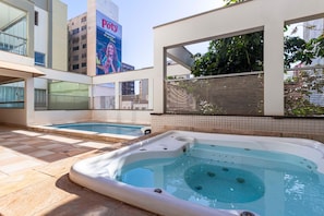 An outdoor swimming pool area with two hot tubs, a tiled floor, and a building with a large billboard in the background.