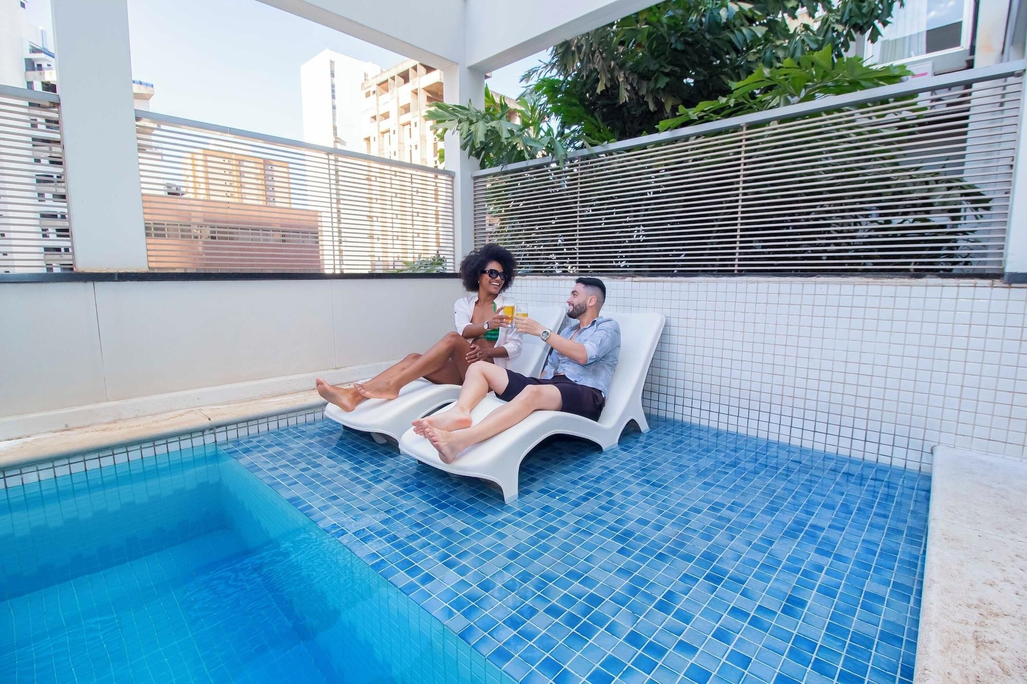 A pool area with two lounge chairs, a tiled floor, and a view of buildings in the background.