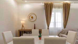 A modern living room with a dining table, white chairs, a sofa, a lamp, a clock, and curtains.