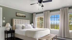 Une chambre avec un grand lit, un ventilateur de plafond, une fenêtre avec des rideaux et une vue sur l’extérieur.