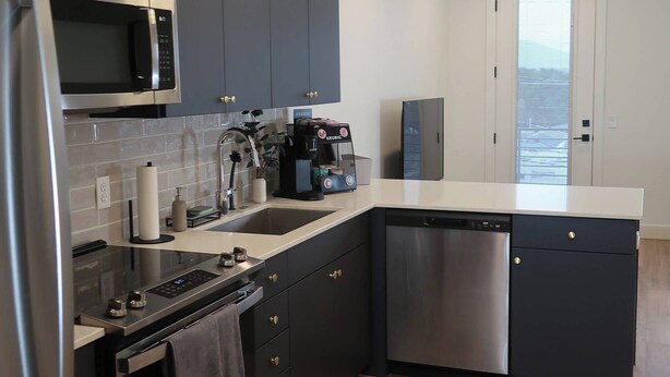 A modern kitchen with dark cabinets, stainless steel appliances, and a large window providing a view of the outdoors. - Cozy studio in marvelous Salt Lake City with WiFi, AC (Salt Lake City)