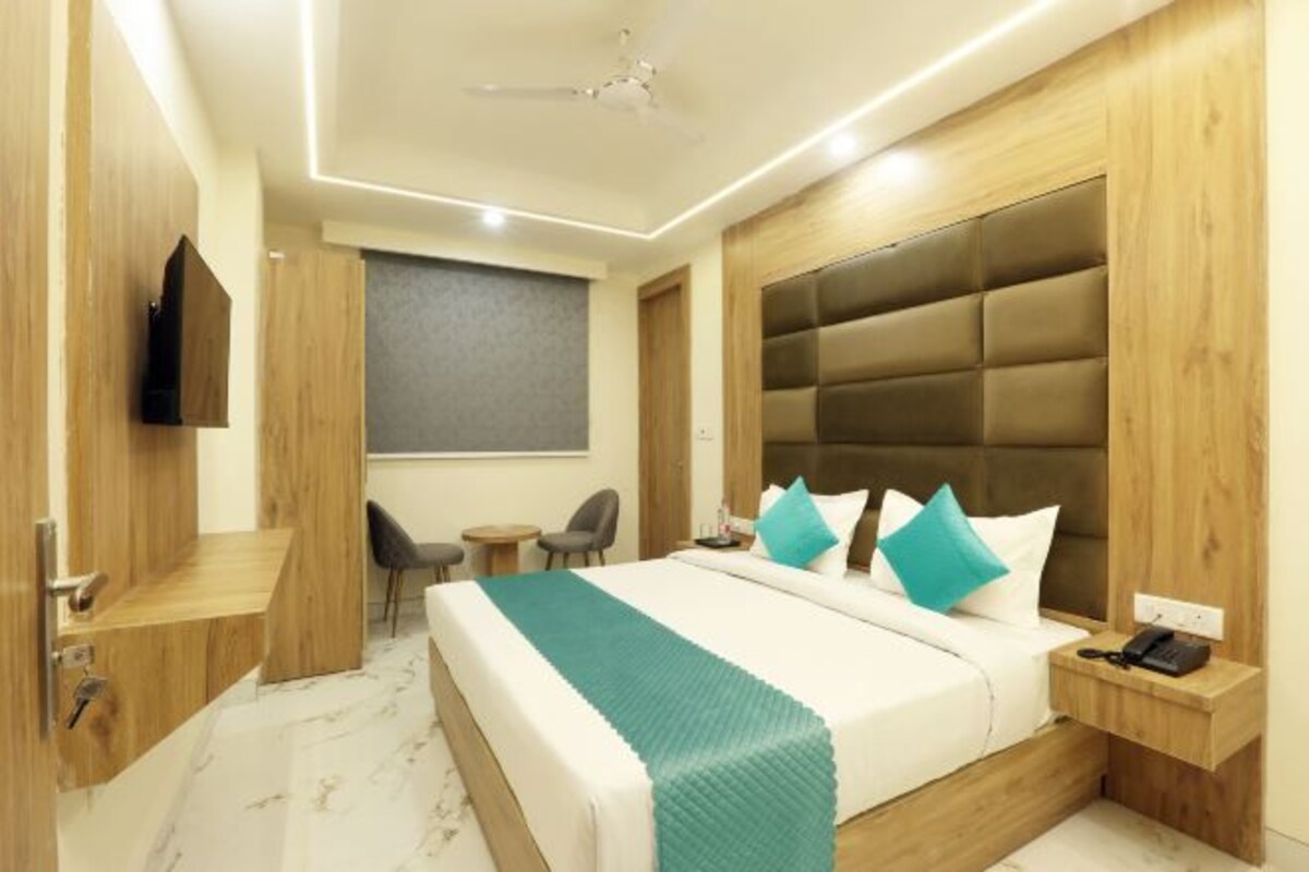 A modern hotel room with a large bed, bedside table, a small desk, a chair, a TV, and a window.