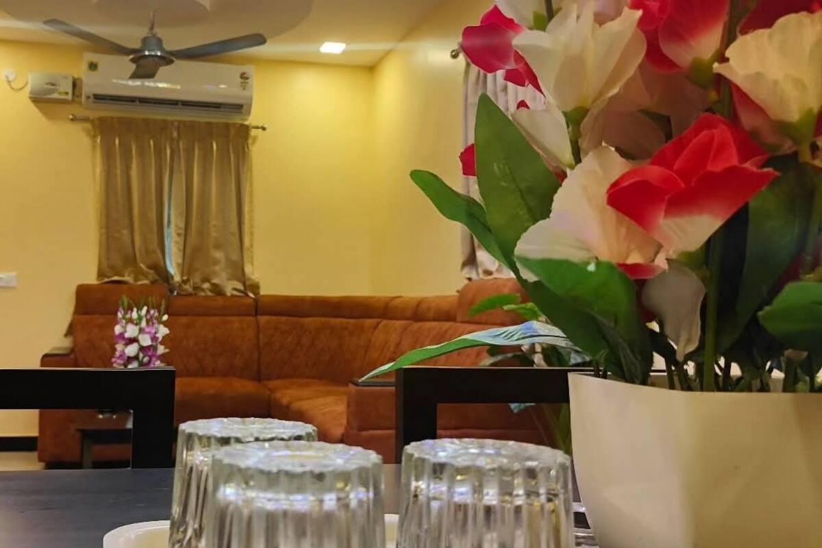 A living room with a brown sofa, a dining table with clear glassware, a ceiling fan, and a wall-mounted air conditioner.