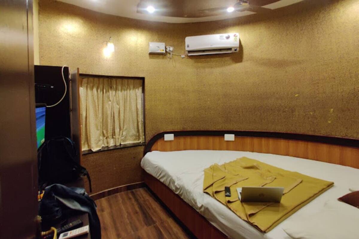 A hotel room with a wooden bed, an air conditioner, a window with curtains, and a desk with a laptop.