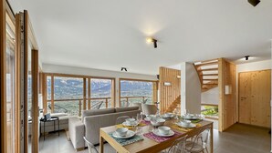 Un espace repas moderne avec une table en bois disposée pour six personnes, entourée de chaises transparentes. De grandes fenêtres offrent une vue sur un paysage montagneux.