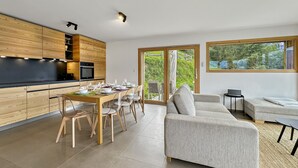 Une cuisine moderne avec des meubles en bois, un coin repas avec une table dressée pour six personnes, et un salon avec un canapé et un ordinateur portable posé sur une table d’appoint.
