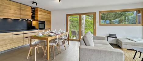Una cocina moderna con armarios de madera, un comedor con una mesa dispuesta para seis personas y una sala de estar con un sofá y una computadora portátil sobre una mesita auxiliar.