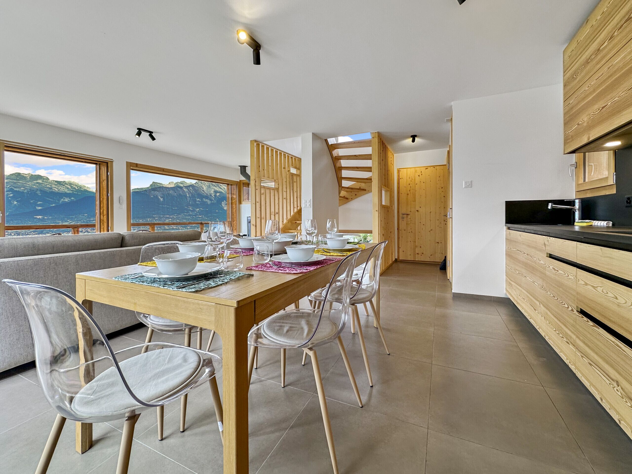 A modern dining area with a wooden table set for six, clear chairs, and a view of mountains through large windows.
