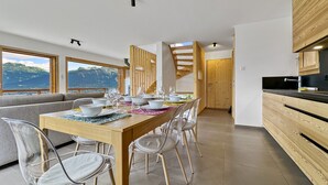 Un espace de repas moderne avec une table en bois dressée pour six personnes, des chaises transparentes et une vue sur les montagnes grâce à de grandes fenêtres.