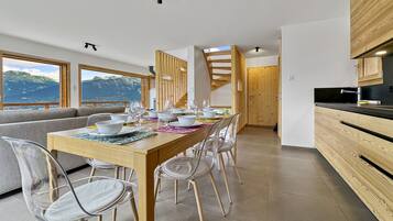 A modern dining area with a wooden table set for six, clear chairs, and a view of mountains through large windows.