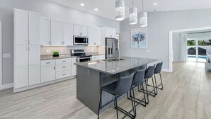 A modern kitchen with white cabinets, a central island with bar stools, and pendant lighting. - Coco Cobana - Heated Pool near Beach & Dining (Naples)