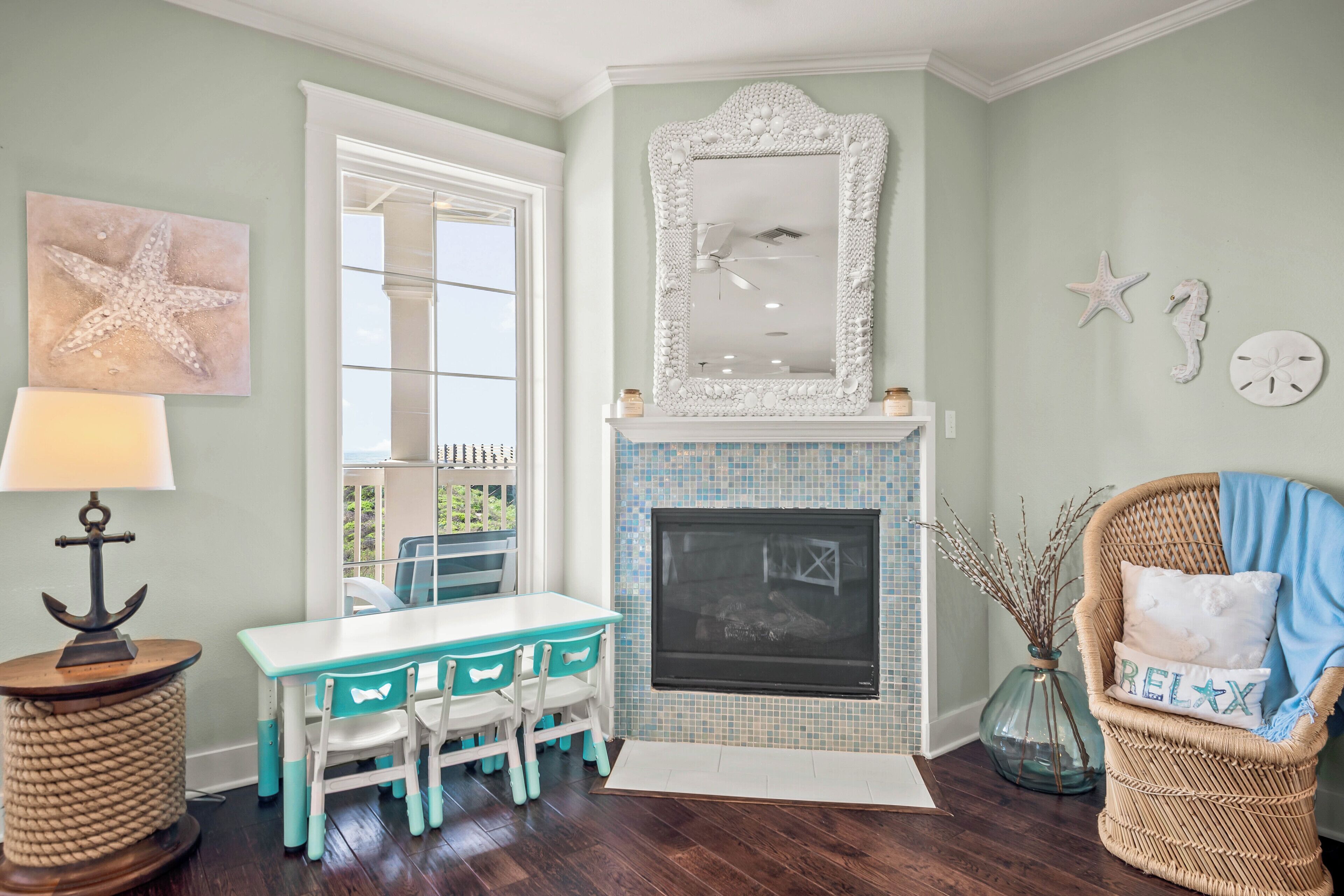 Quarto com lareira branca, cadeiras cor verde-água, uma cadeira de vime com almofada 'RELAX' e decoração de parede com estrela-do-mar.