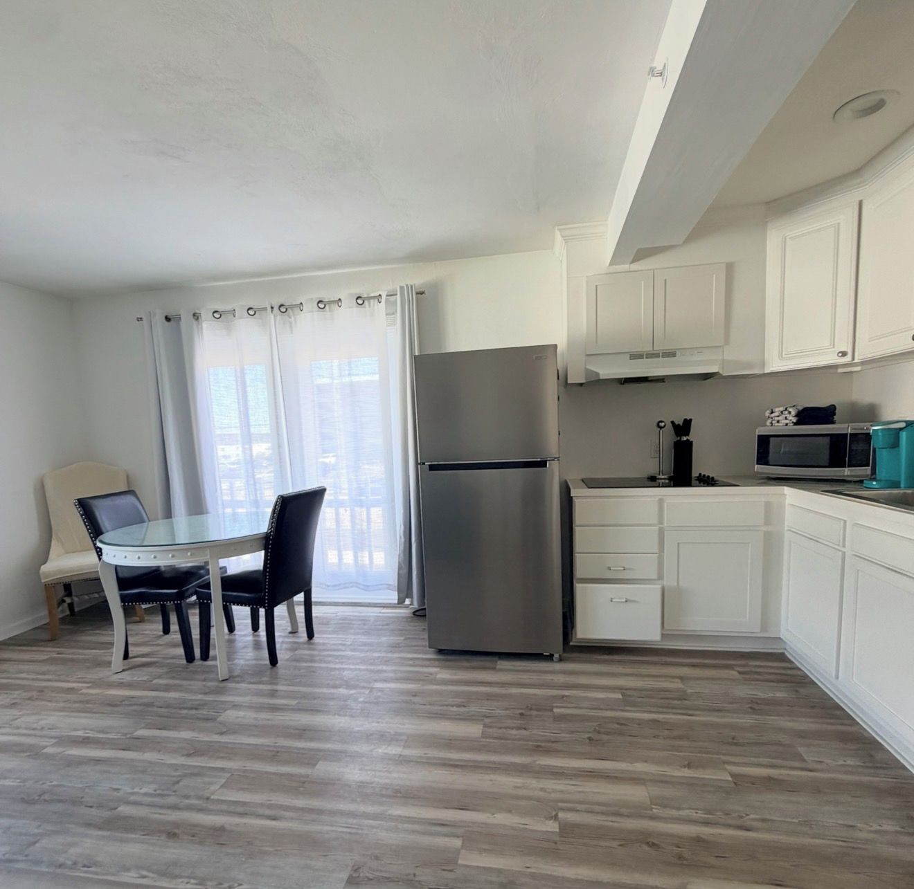 A modern kitchen with white cabinets, a stainless steel refrigerator, a microwave, and a dining area with a glass table and chairs.