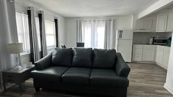 A living room with a dark leather sofa, a small side table, and a lamp. There is a kitchen area with white cabinets and a microwave.