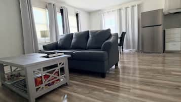 A modern living room with a grey sofa, a coffee table, and a kitchen area with a stainless steel refrigerator.