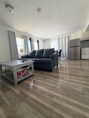 A modern living room with a grey sofa, a coffee table, and a kitchen area with a stainless steel refrigerator.