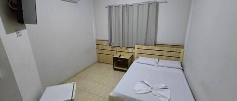 A small, clean room with a bed, a bedside table, a nightstand, a TV, and an air conditioning unit.