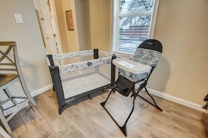 A room with a wooden floor, a crib with a patterned mattress, a high chair, and a window with blinds.