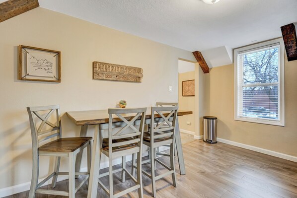A dining area with a wooden table and four chairs, a framed artwork, a sign, and a window with blinds.