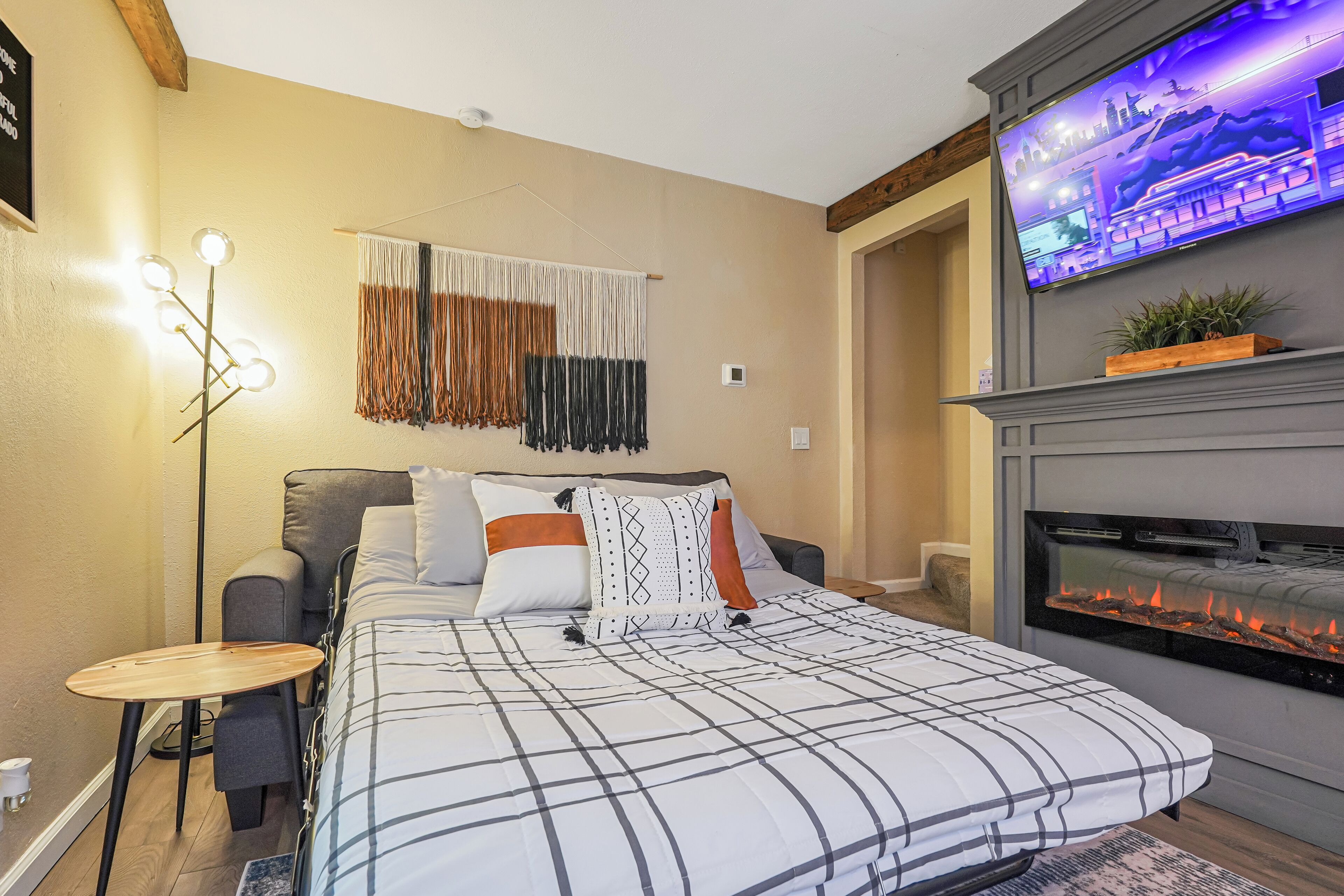 A bedroom with a bed, a TV mounted above a fireplace, and a small table with a lamp.