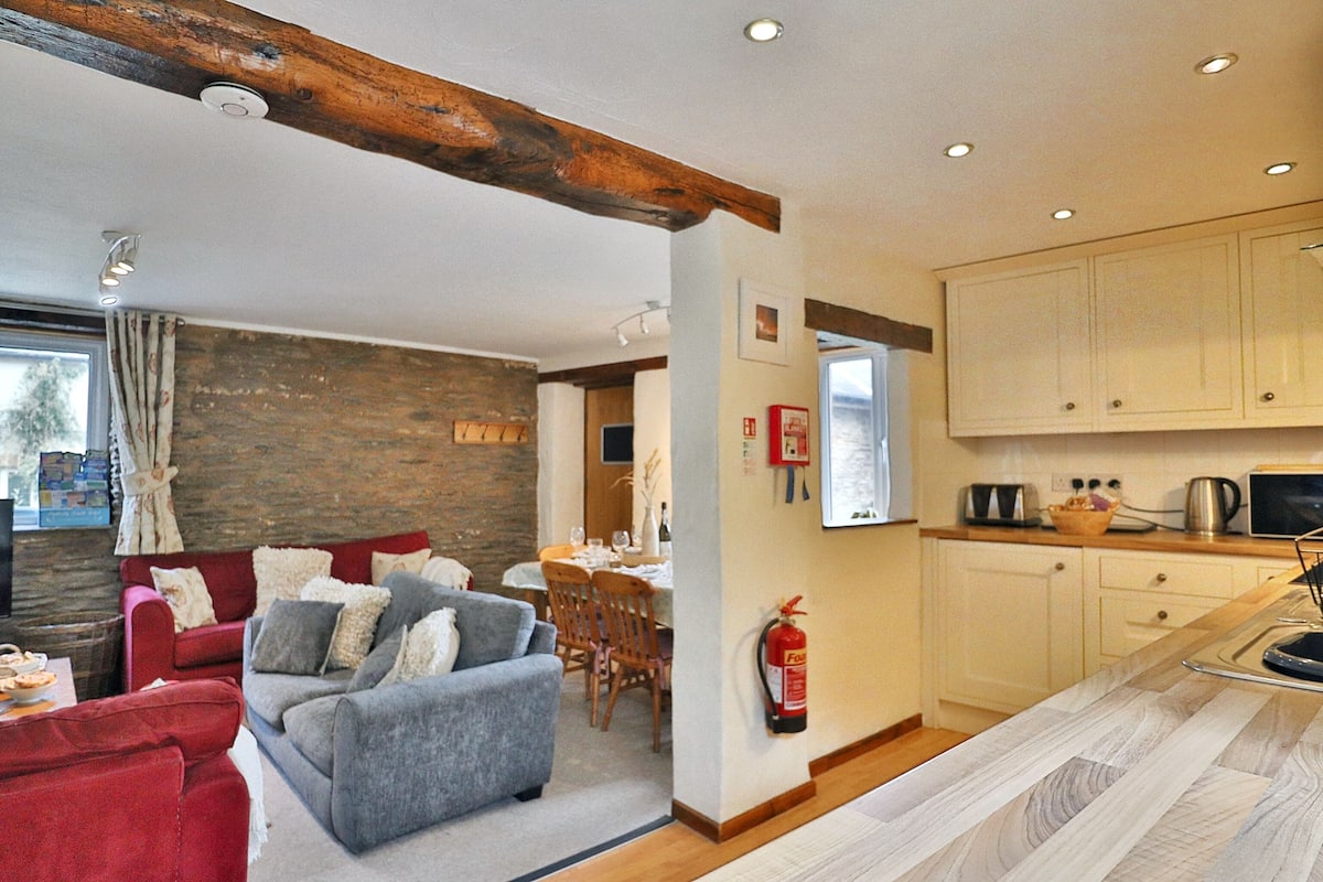 Ein modernes Wohnzimmer mit grauem Sofa, einem roten Sessel und Holzbalken an der Decke. Im Hintergrund ist ein Küchenbereich mit Holzausbau und einem Esstisch zu sehen.