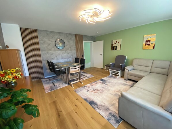 A modern living room with a dining area, a grey sofa, a grey armchair, a dining table with chairs, a wooden cabinet, and a potted plant. - Candel view (Waldkirch)