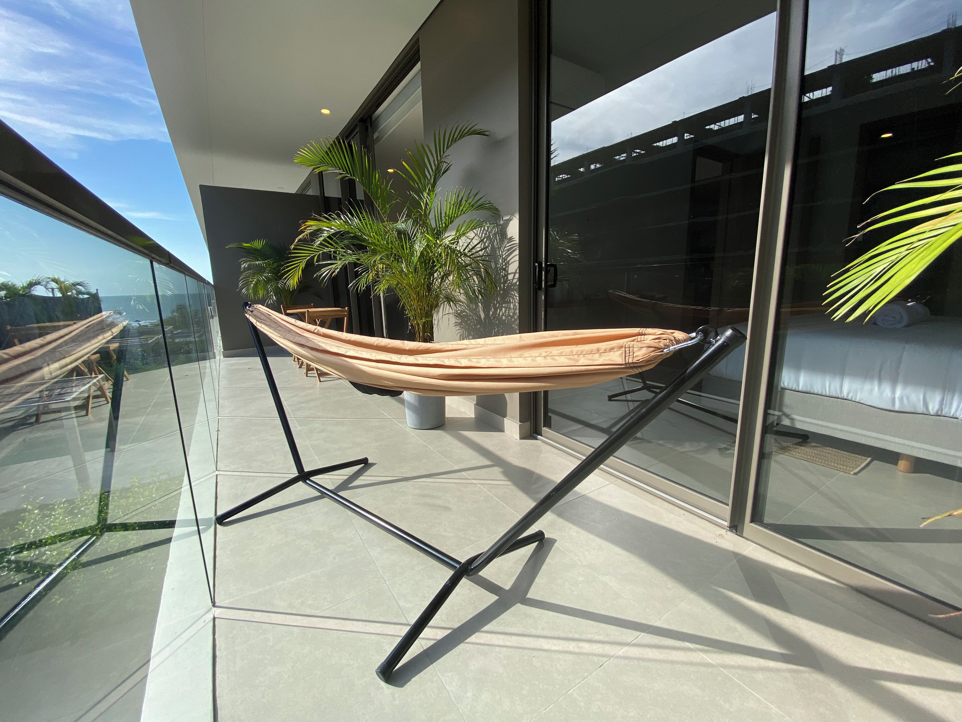A hammock on a balcony with a glass railing, a potted plant, and a view of a building.