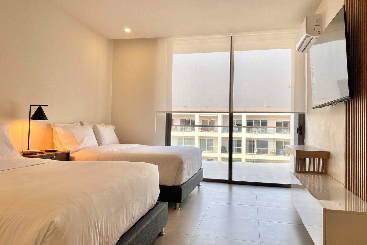 A hotel room with two beds, a nightstand, a lamp, a TV, and a balcony with a view of other buildings.