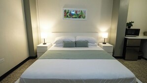 Siististi järjestetty hotellihuone, jossa on sänky, kaksi yöpöytävalaisinta, pieni pöytä, jossa on kasvi, ja mikroaaltouuni.