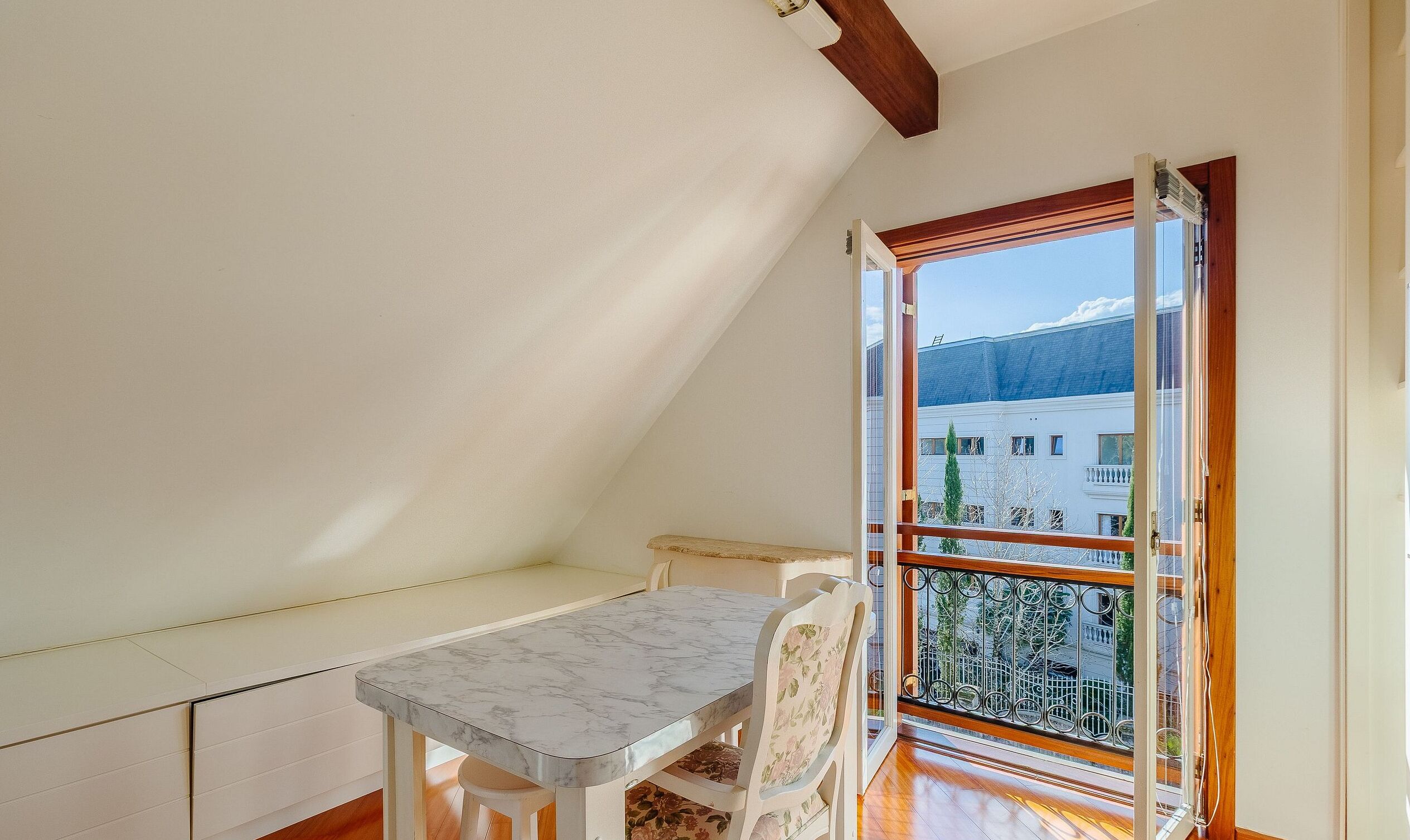 A small balcony with a table and chairs, a view of a building, and a wooden ceiling beam.
