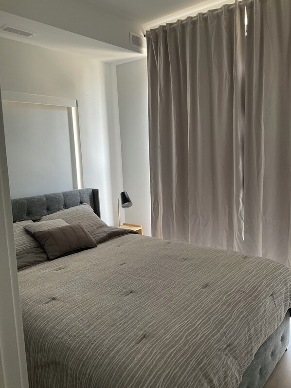 Ein Schlafzimmer mit einem Bett, einem Nachttisch, einer Lampe und einem Fenster mit Vorhängen.