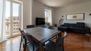 En moderne stue med spisebord, stole, sofa, fjernsyn og en altan med udsigt til bygninger.