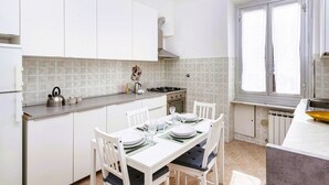 Une cuisine avec des meubles blancs, une table à manger dressée pour quatre personnes, une cuisinière et une fenêtre avec des stores.
